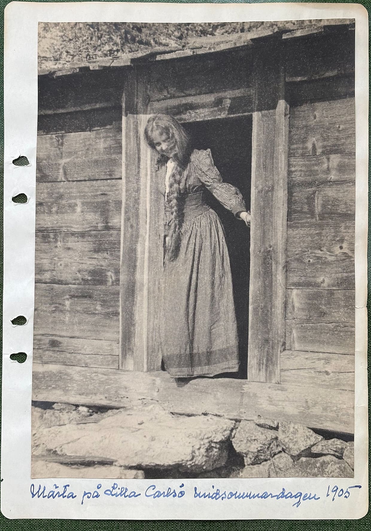 Fotograferad dagbokssida 135. Helsidesfotografi: en ung kvinna med lång fläta står i dörröppningen till en timrad bod, klädd i randig kjol och blus, med handen på dörrposten. Bildtext: 'Märta på Lilla Carlsö midsommardagen 1905'.