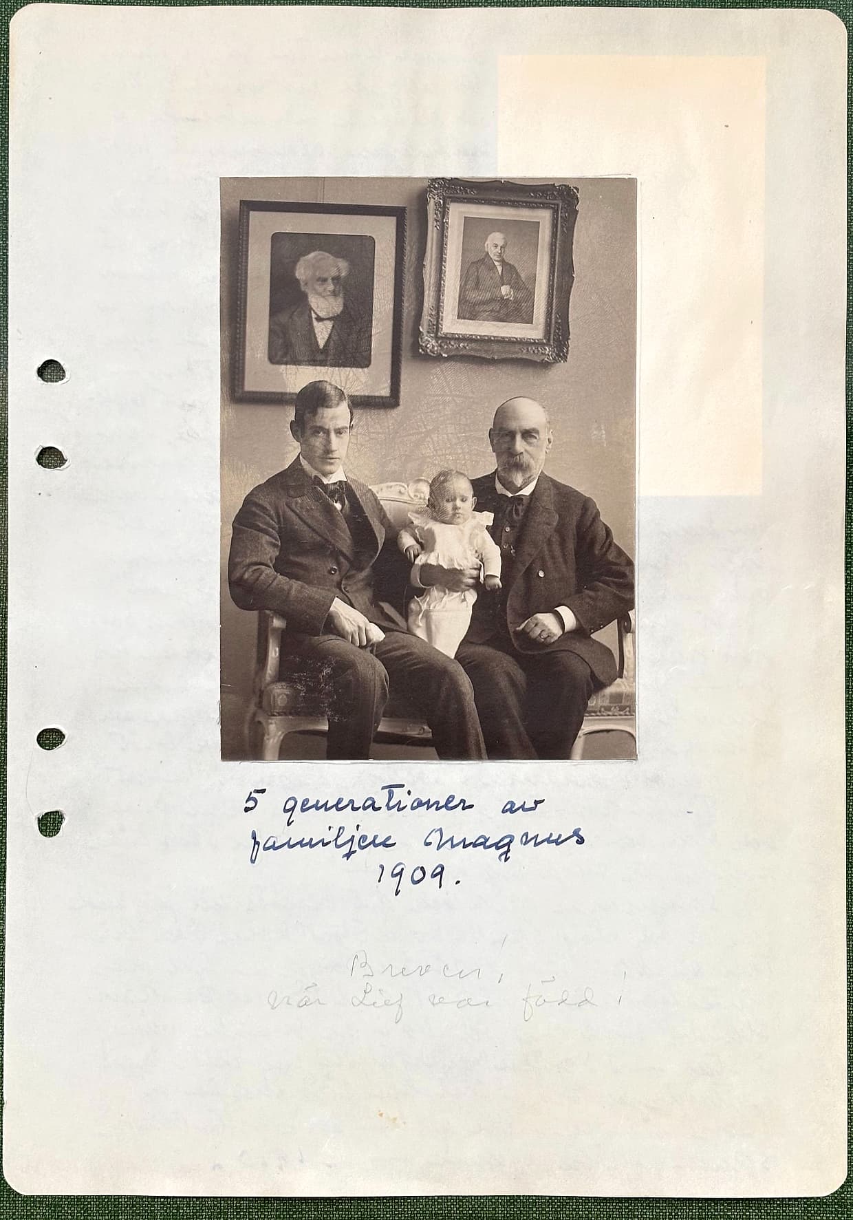 Fotograferad dagbokssida 169. Foto av två män med ett spädbarn mellan sig, framför en vägg med två inramade porträtt. Bildtext: '5 generationer av familjen Magnus 1909.'.
